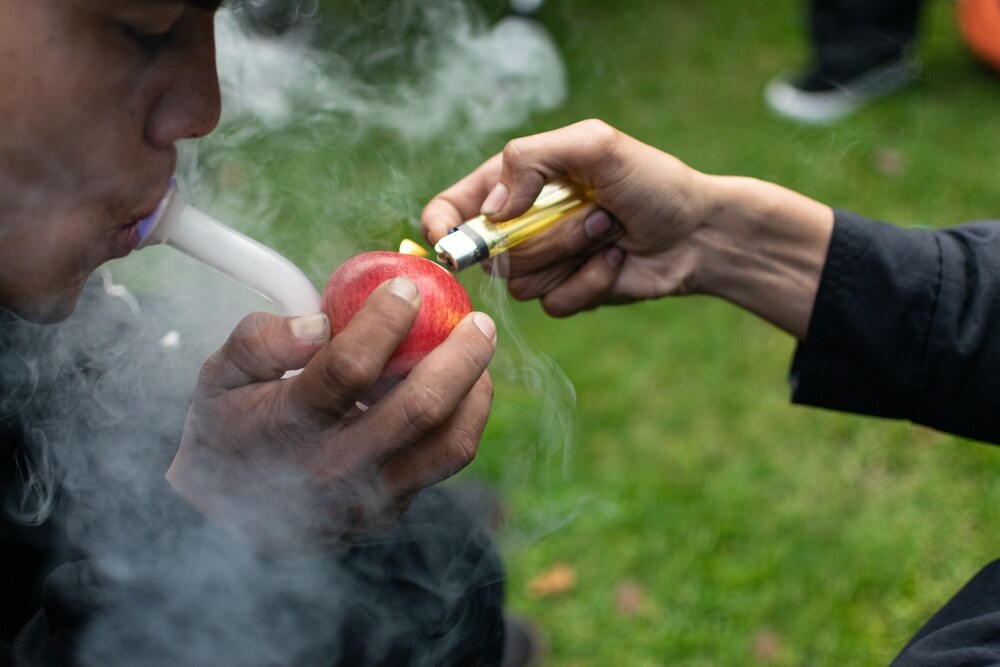 a man smoking from a pipe connected to an apple with his friend helping him burn the bud
