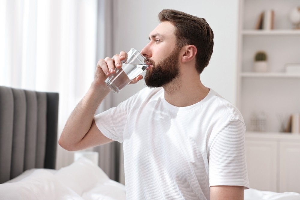 a man in a white t-shirt drinking water out of a glass in his bedroom