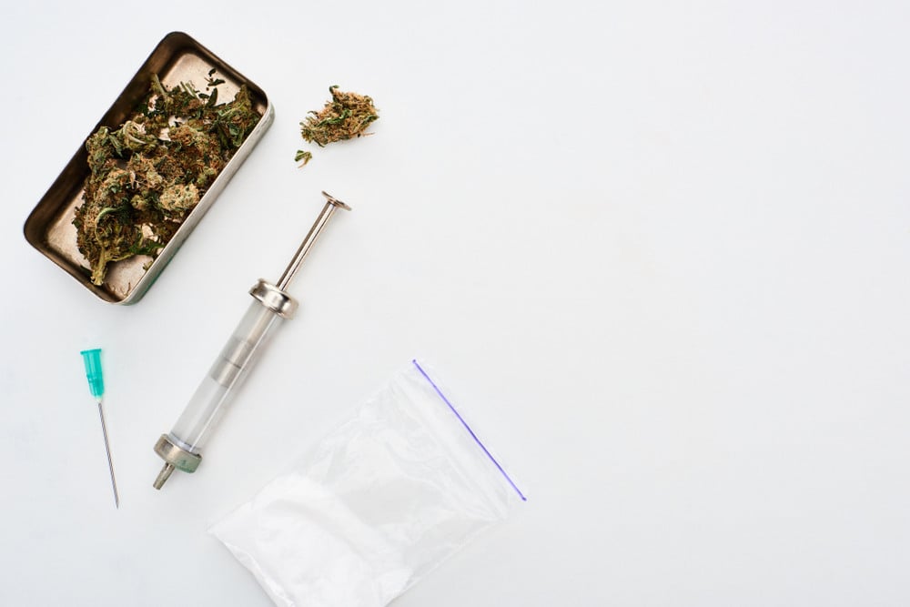 cannabis bud in a silver tray with a needle and a baggie of a white substance on a white table