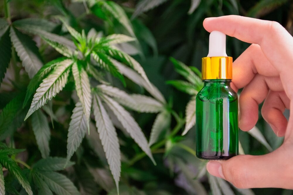 a man holding up a green bottle of cannabis oil next to a cannabis plant