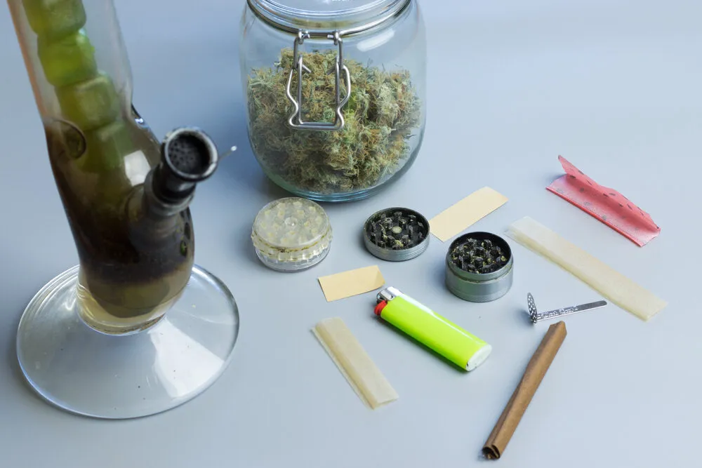 different accessories to use for smoking cannabis including a grinder, papers and a lighter with a jar of cannabis bud at the back