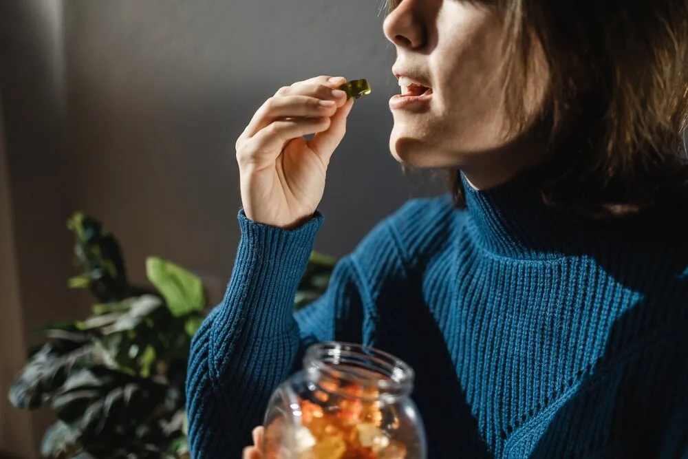 a lady in a blue jumper eating cannabis gummy edibles from a jar with a house plant in the background