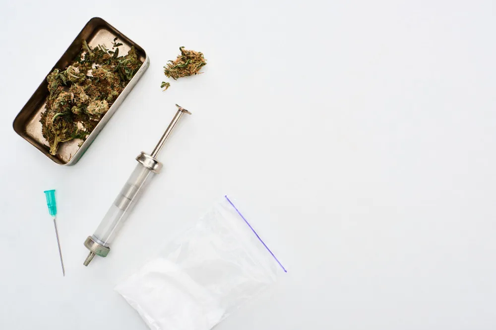 cannabis bud in a silver tray with a needle and a baggie of a white substance on a white table