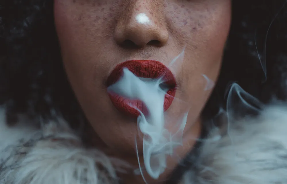 a lady with red lipstick, freckles and a fur coat blowing out smoke from cannabis intake