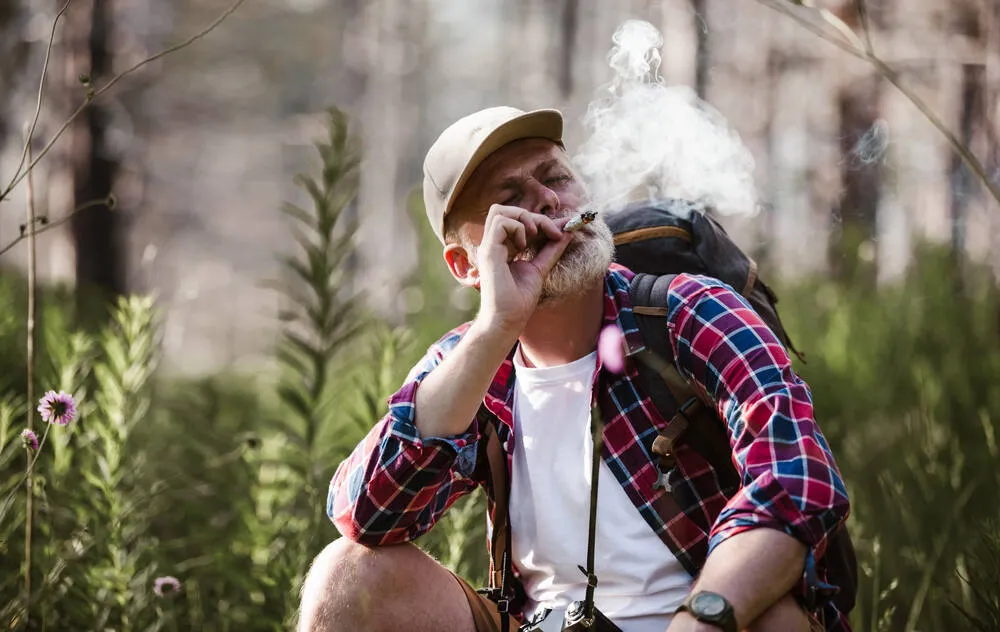 a man in a chequered shirt in the forest taking a draw from a cannabis joint